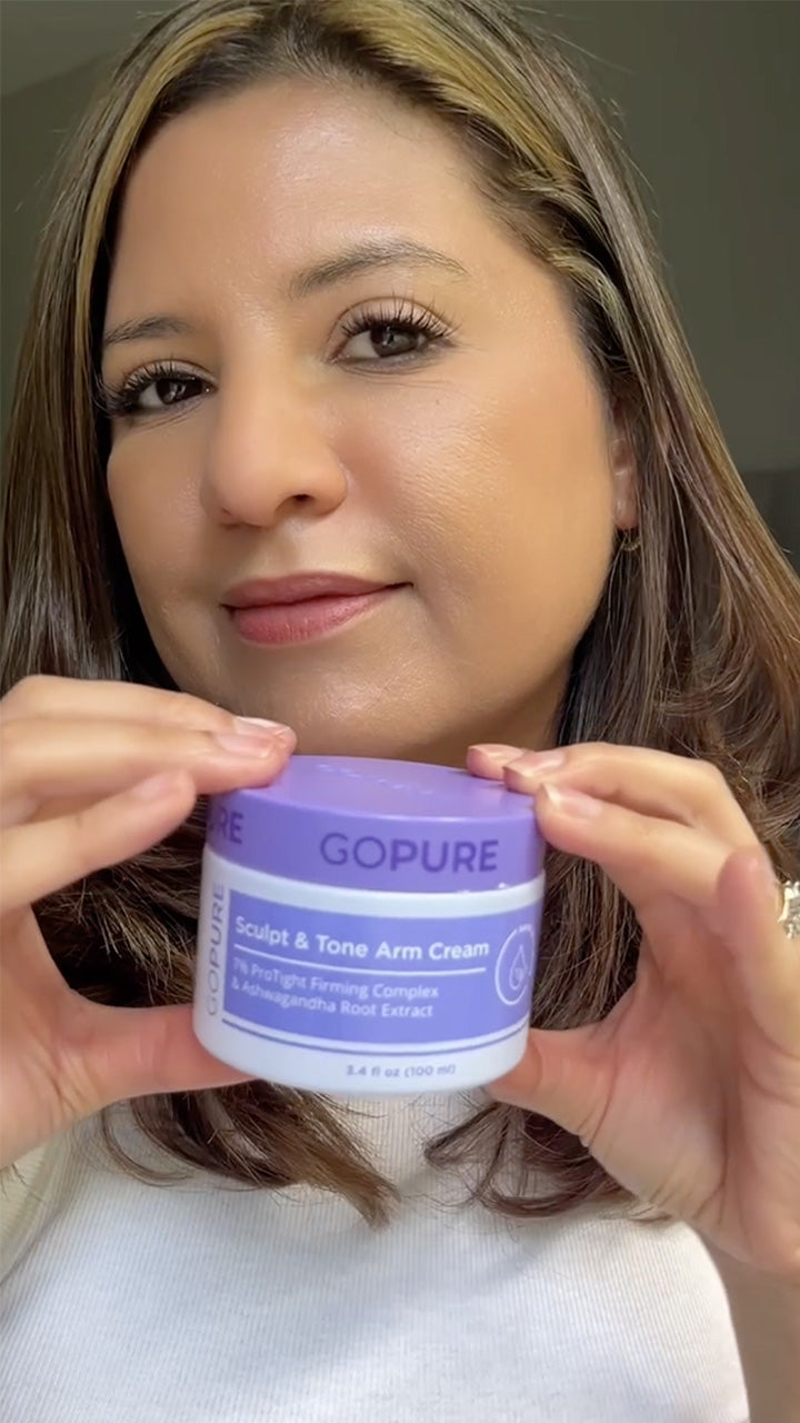 A woman with light brown hair holds up a jar of “GOPURE Sculpt & Tone Arm Cream” close to the camera, displaying the product label. She has a natural, gentle smile and is wearing a light top, with soft lighting in the background.