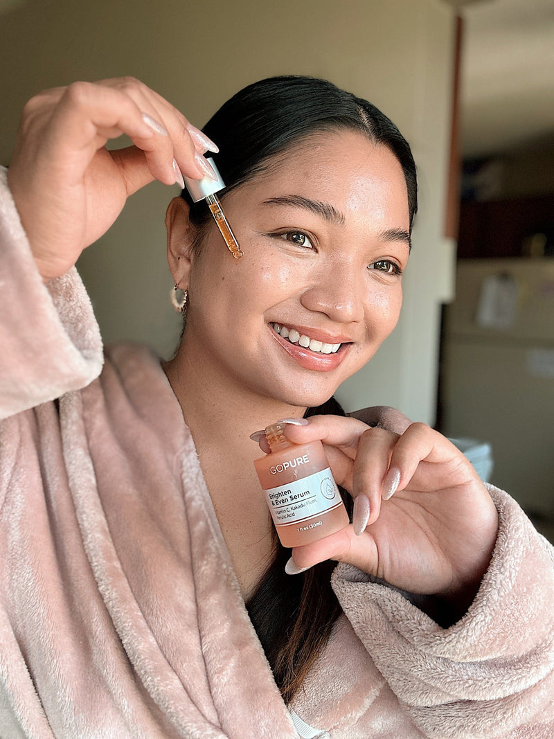 Smiling woman applying GoPure Firm & Lift Serum to her face using a dropper. She is wearing a light pink robe and is posed in a cozy indoor setting.