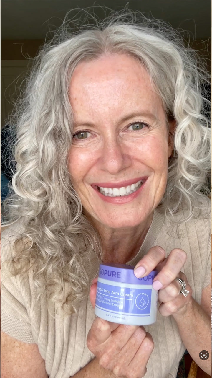 A woman with curly gray hair and a warm smile holds a jar of “GOPURE Sculpt & Tone Arm Cream” in both hands, positioned close to her face. She is wearing a beige sweater, and the background is softly lit.