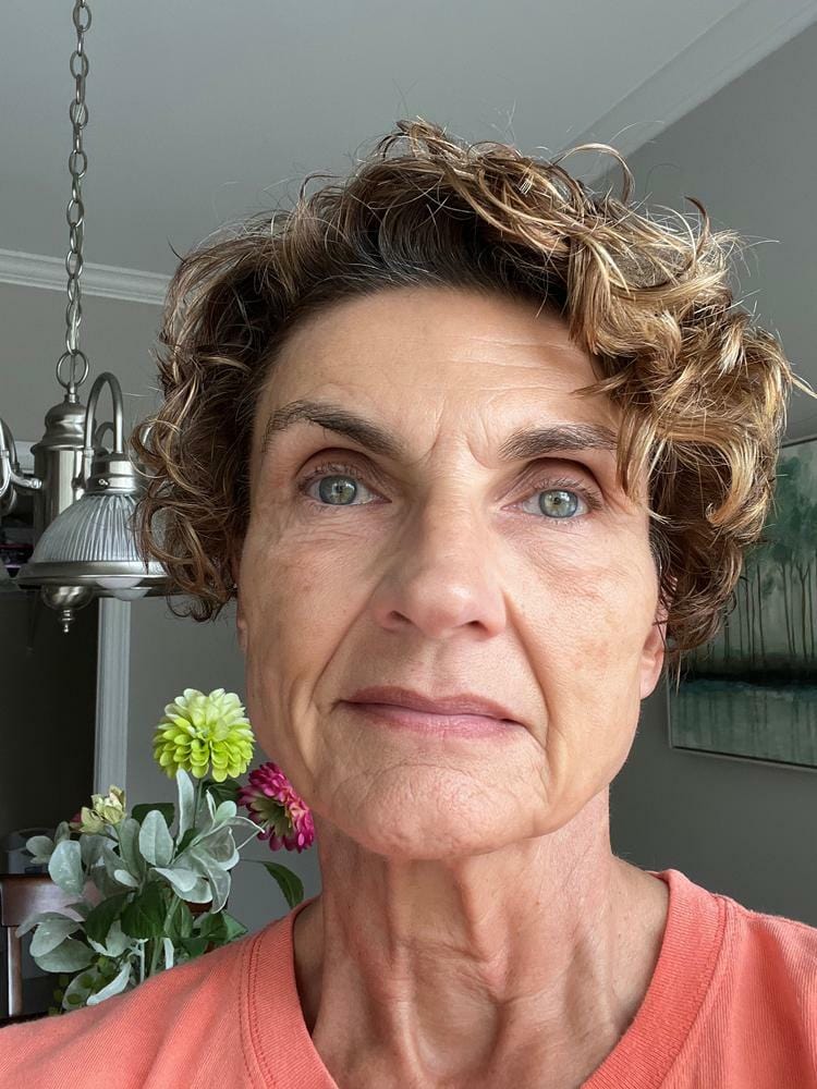 A woman with short, curly hair taking a selfie indoors, wearing a coral-colored top. The background includes a vase of colorful flowers and a modern light fixture, adding a warm and inviting atmosphere. Ideal for topics like 'natural portrait' or 'indoor photography.