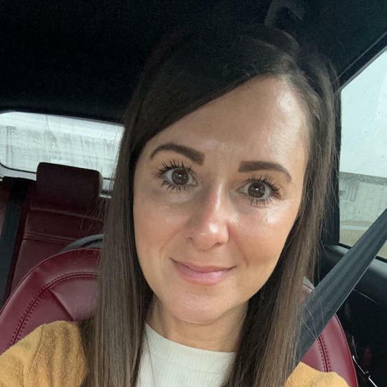 A woman with long, straight hair taking a selfie in a car, wearing a seatbelt and a neutral expression. The car interior features red leather seats