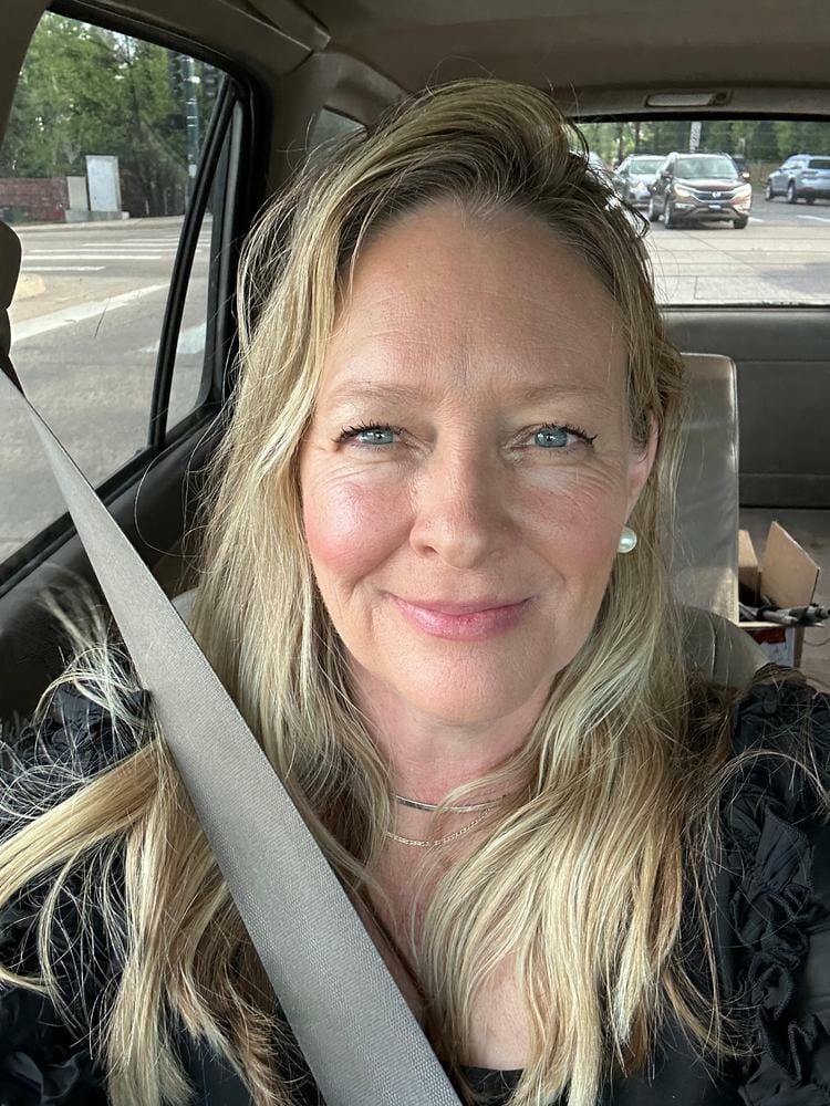 A woman taking a natural selfie in a car while wearing a seatbelt, with a slight smile and natural lighting. The background shows a street with cars and trees, enhancing the candid and relatable vibe. Perfect for themes like 'woman driving' or 'natural selfie.