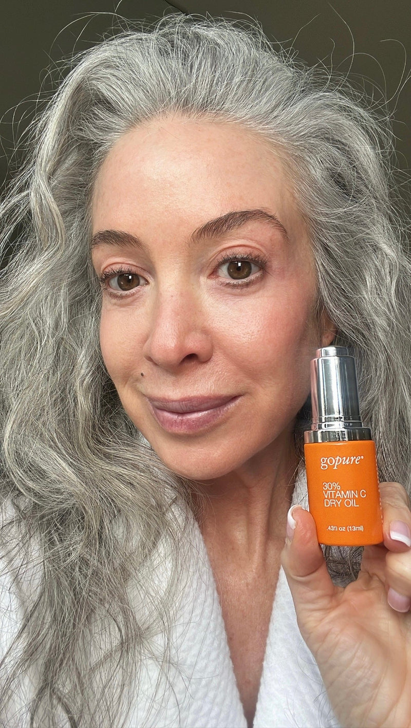  Woman with long gray hair holding a bottle of GoPure 30% Vitamin C Dry Oil. She is smiling and posed against a dark background.